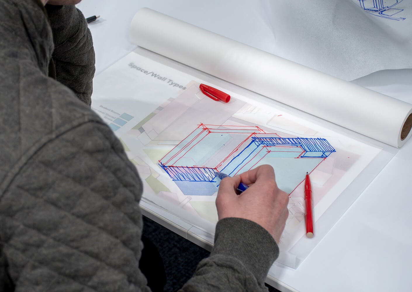Person sketching architectural designs in blue and red on transparent paper.
