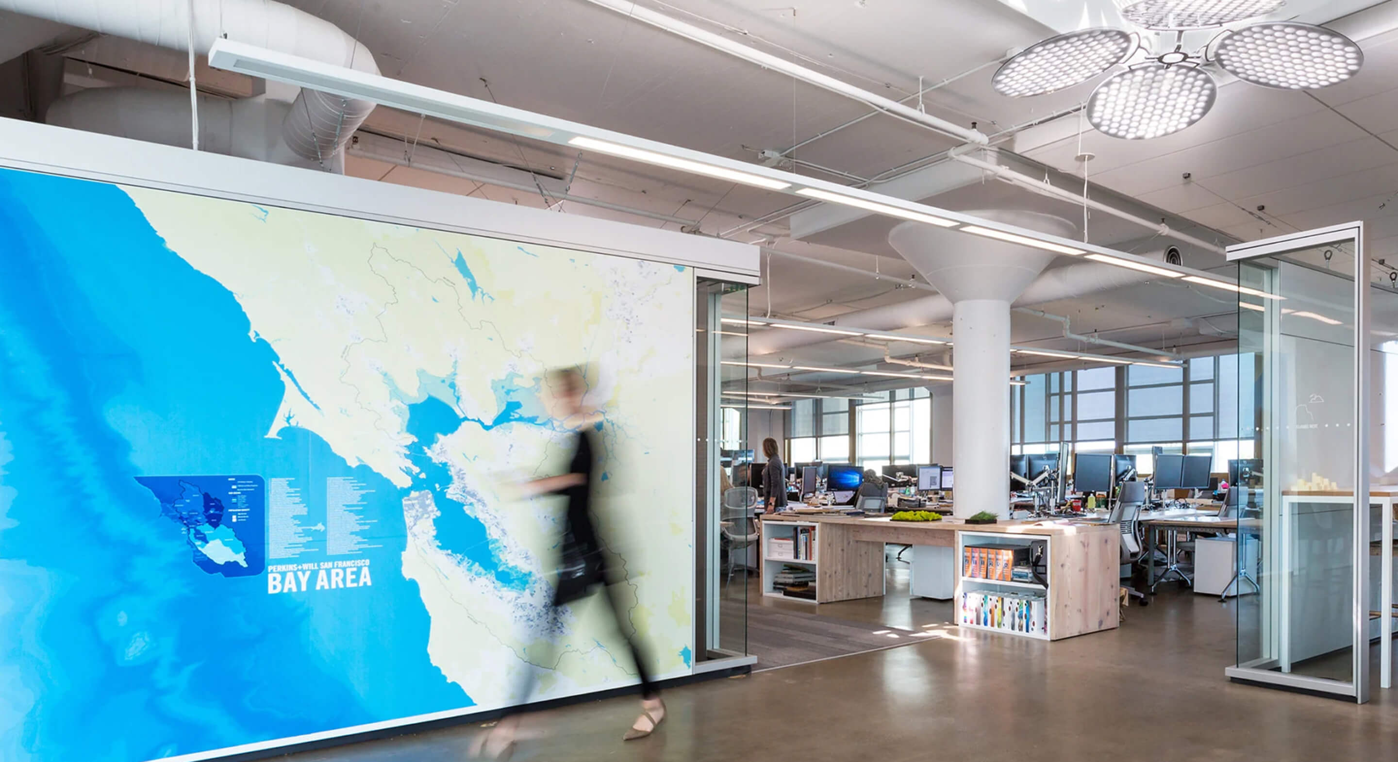 Office space featuring a large Bay Area map on the wall, with employees working at desks and a person walking past. Bright, industrial design elements visible.