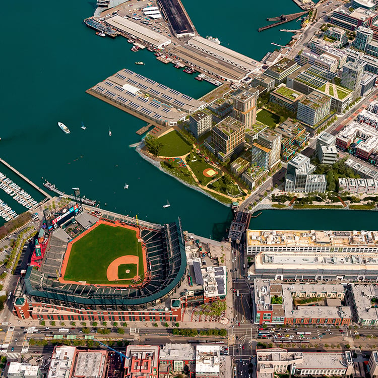 Aerial view of a waterfront area featuring a baseball stadium, residential buildings, a marina, and landscaped parks along the water's edge.