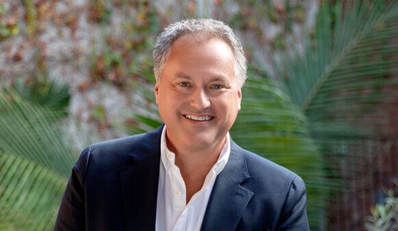 Headshot of a smiling man in a dark blazer and white shirt, surrounded by green plants, against a textured background.