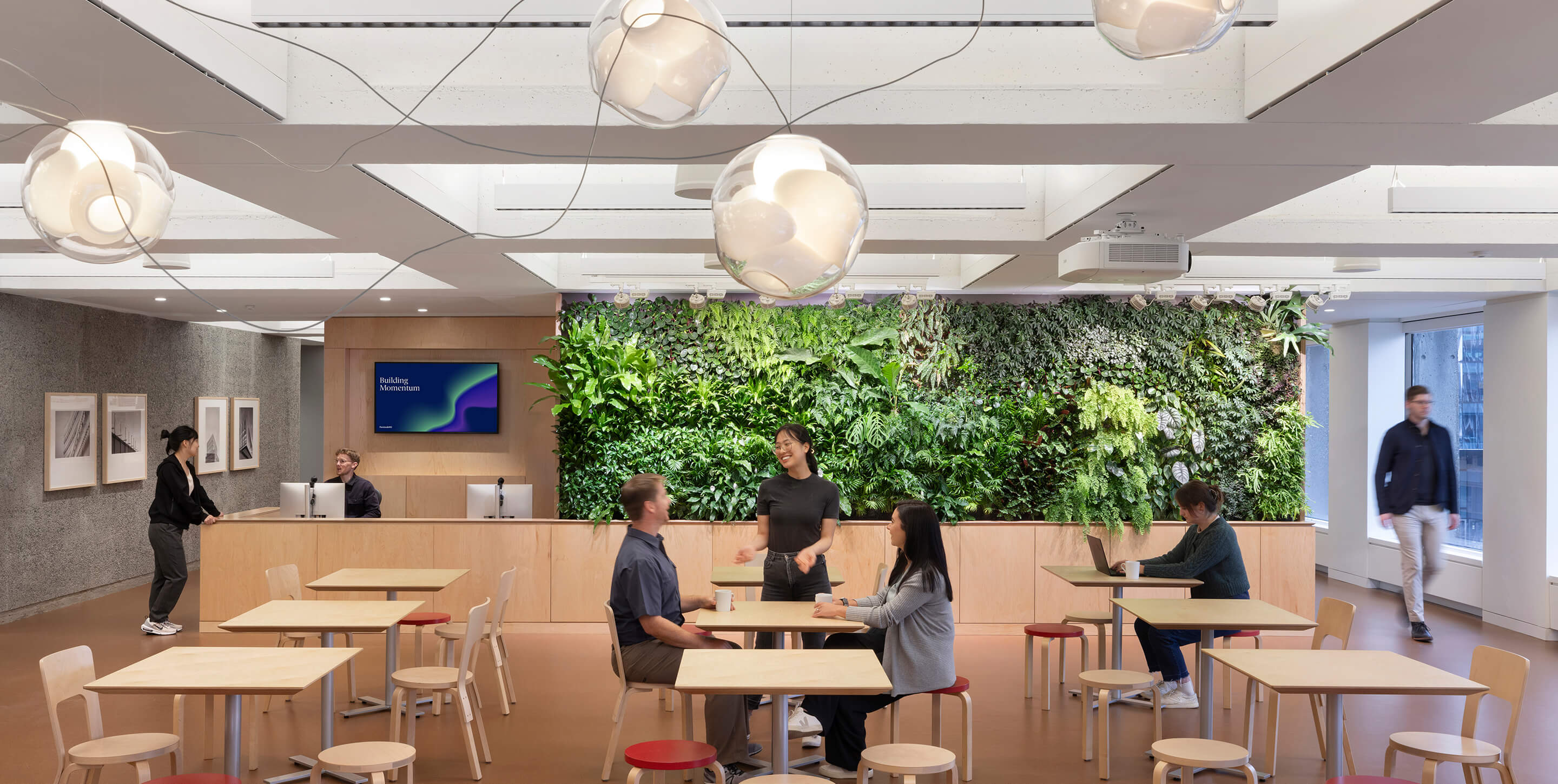 Studio cafe with reception and a green wall, seating, and individuals interacting. Light fixtures hang from the ceiling, enhancing workspace ambiance.
