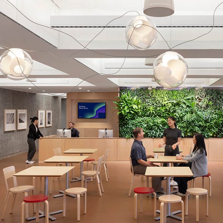 Studio cafe with reception and a green wall, seating, and individuals interacting. Light fixtures hang from the ceiling, enhancing workspace ambiance.