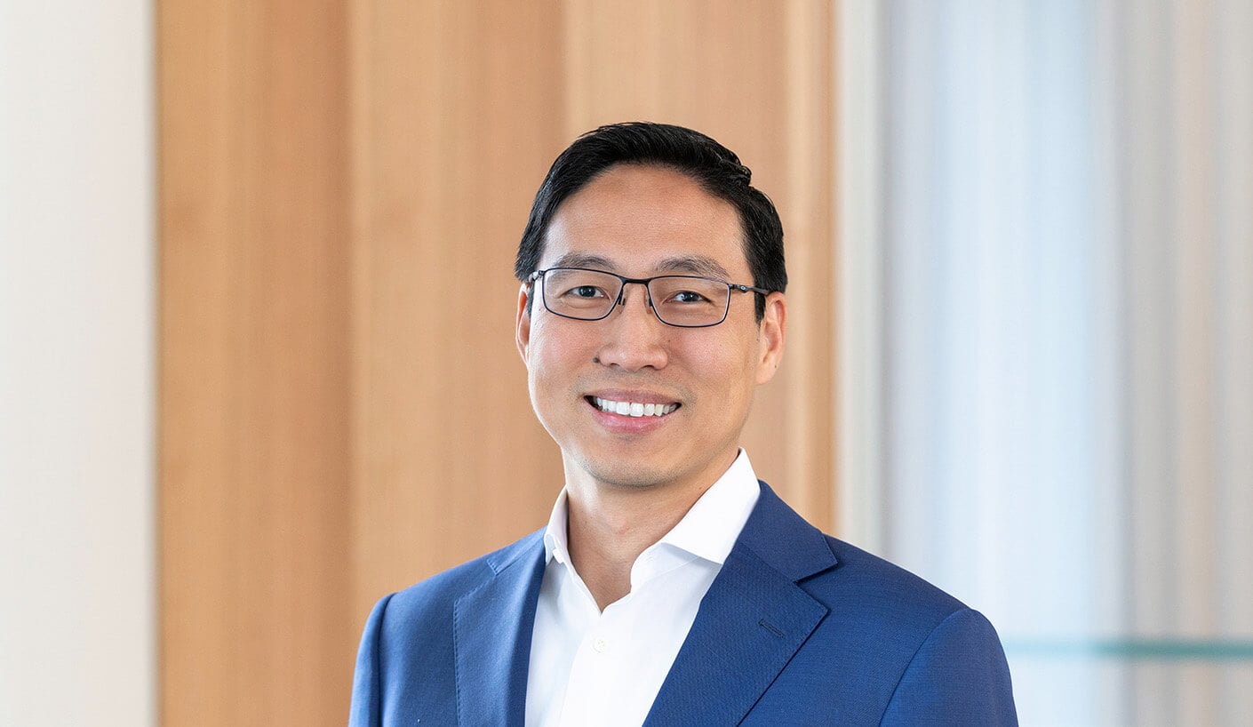 Business professional with glasses wearing a blue suit, smiling in an office setting with wooden panels in the background.