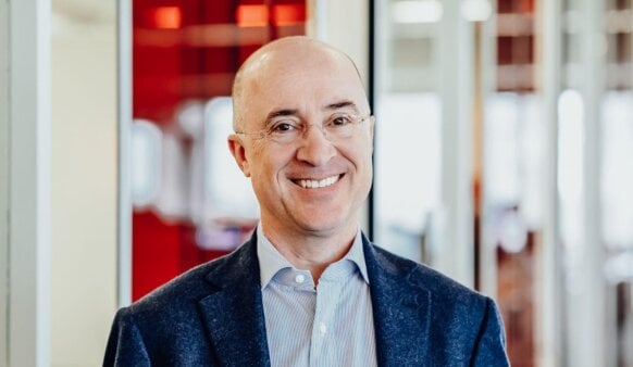 Smiling professional man in blue blazer, glasses, standing in modern office with red accents.