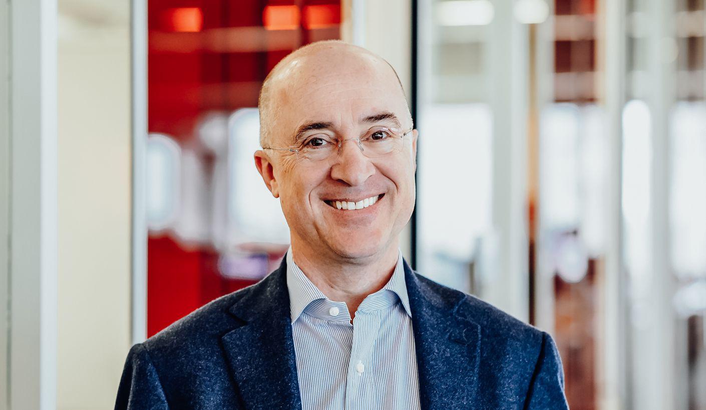 Smiling professional man in blue blazer, glasses, standing in modern office with red accents.