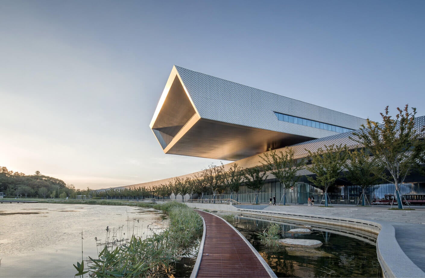 Contemporary architectural building overlooking a tranquil waterway, featuring angular design and surrounding landscaped areas with pathways.