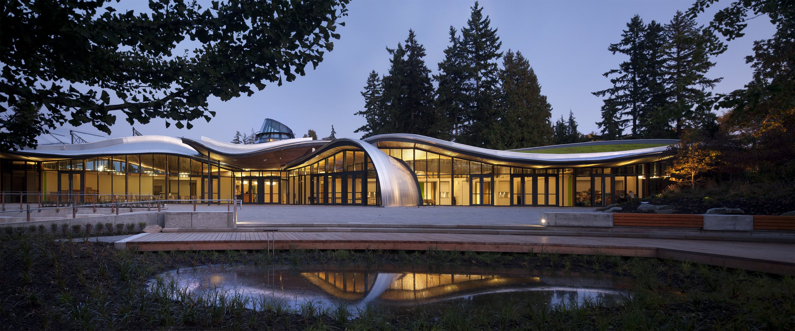 Modern building with curved roof and glass walls, surrounded by trees and reflecting pond.