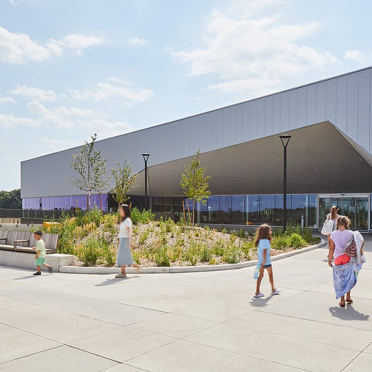 Modern building exterior with people walking by landscaped entrance on a sunny day.