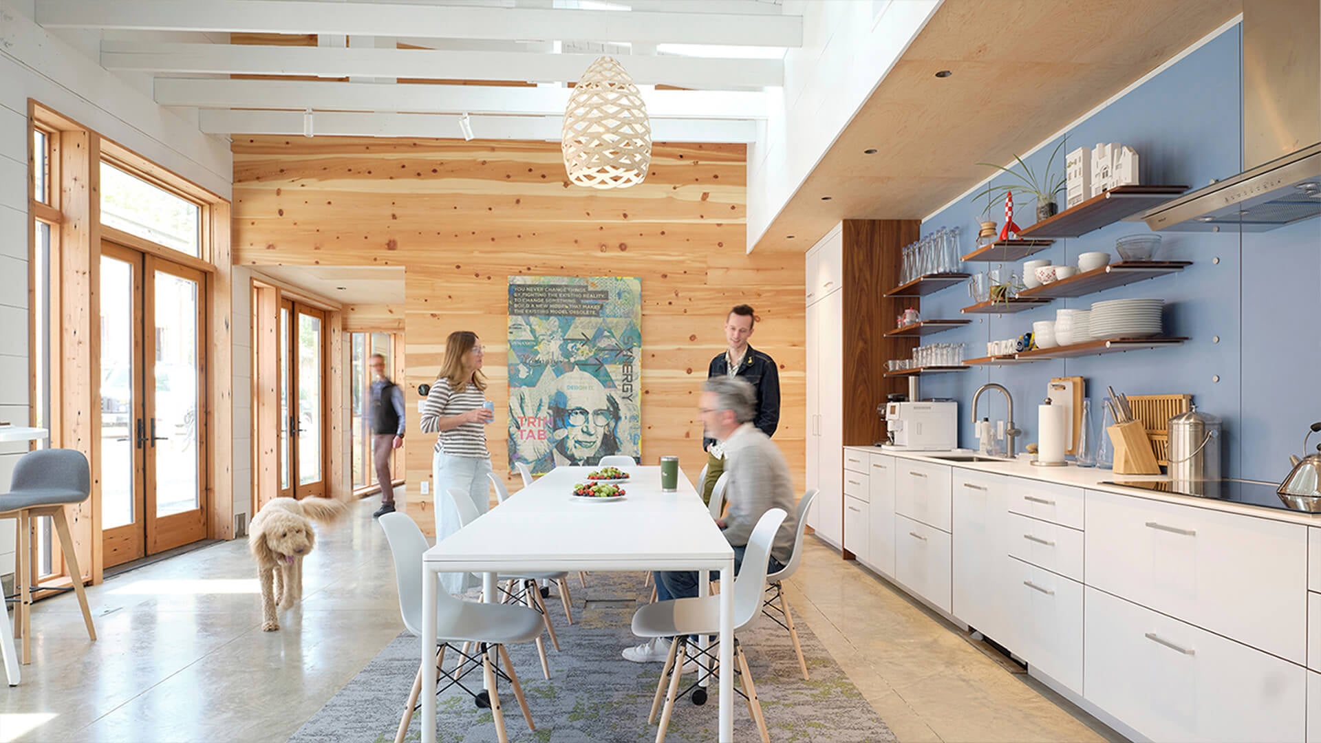 Bright kitchen and dining area with wooden accents, featuring a dining table, people interacting, and a dog walking by. Natural light flows in through large windows.