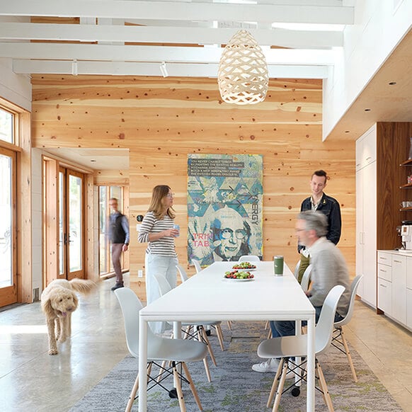 Bright dining area with large table, wooden walls, and a hanging light. Two men and a woman engage while a dog walks through.