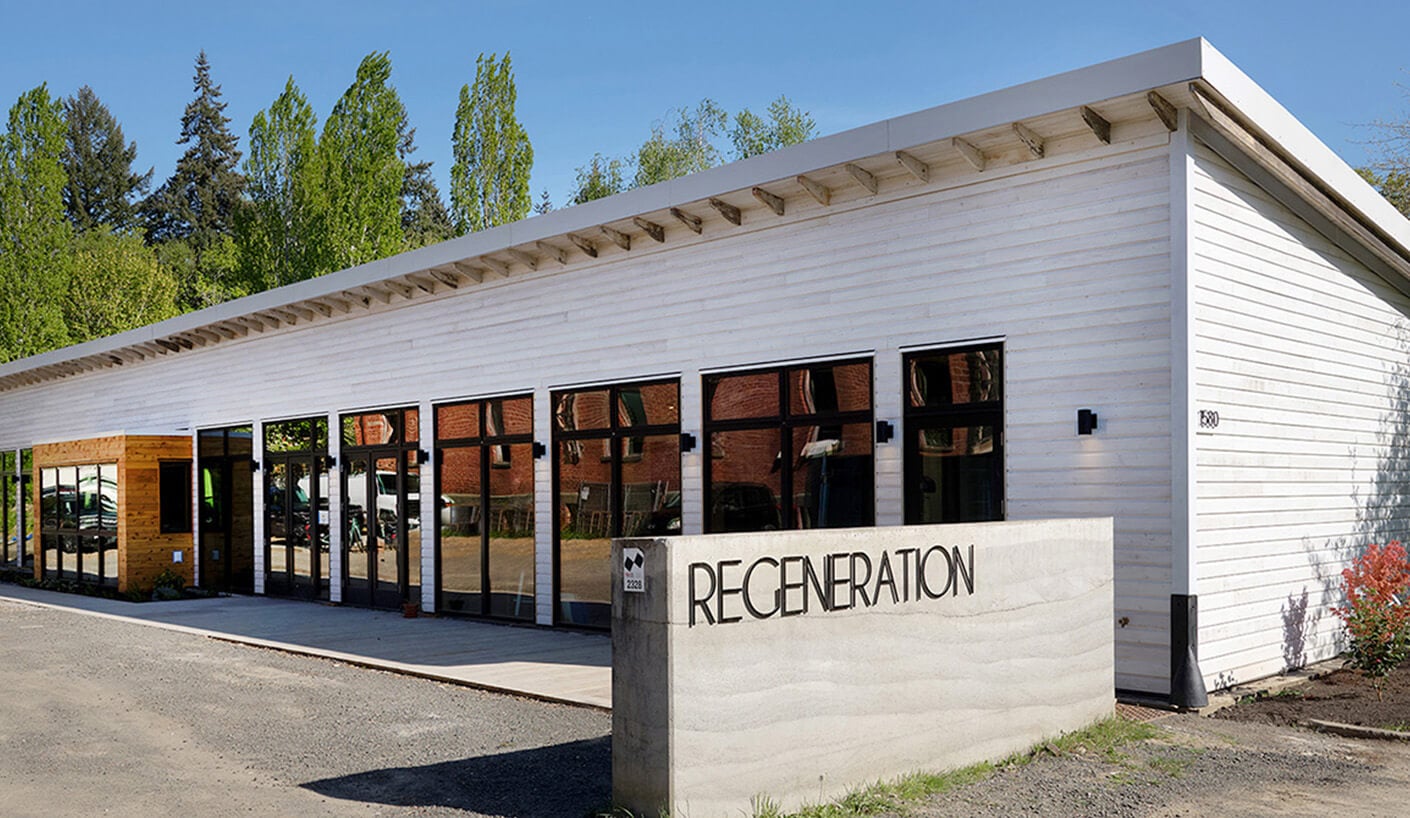 Exterior view of the Regeneration building, featuring large windows, a wooden facade, and landscaping, under a clear blue sky.