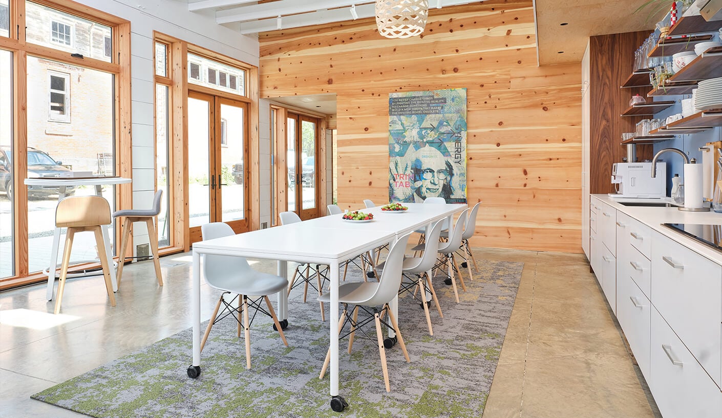 Bright dining area featuring a long white table, gray chairs, large windows, and wooden walls. A colorful artwork is displayed nearby.