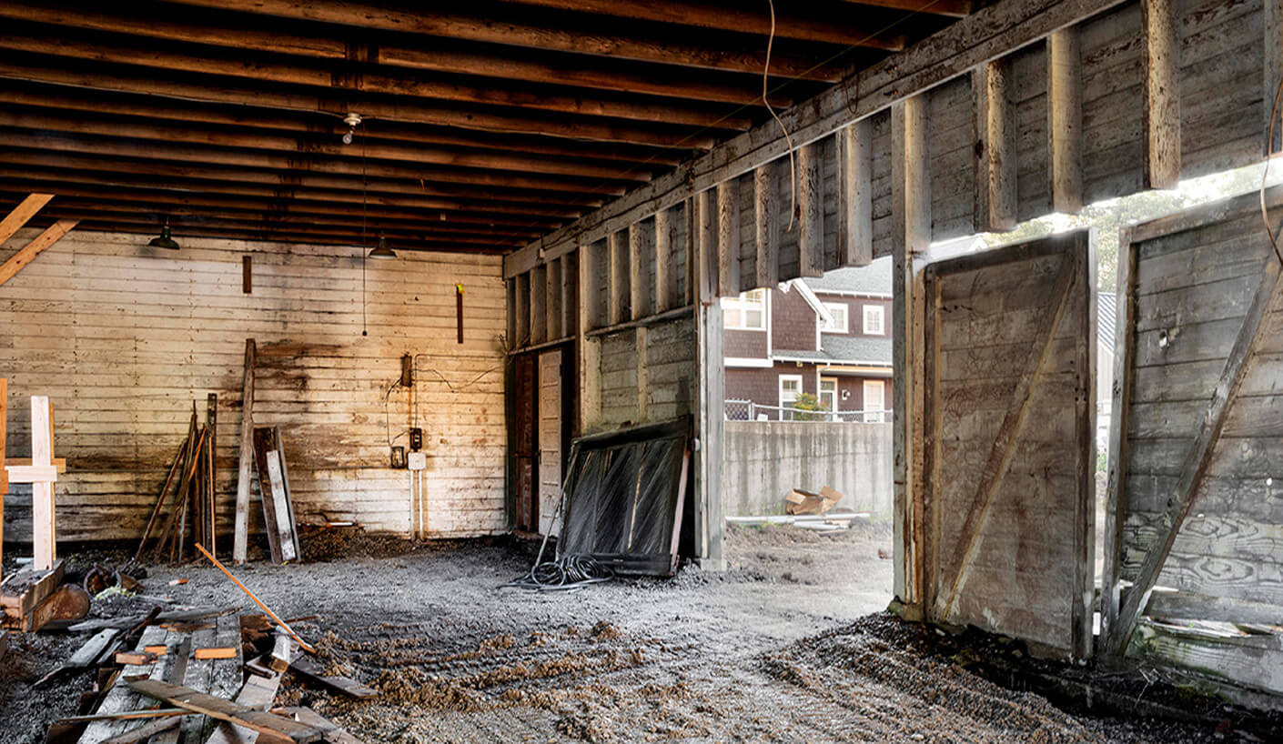 Interior of a vacant space with exposed wooden beams, dirt floor, and construction materials stacked against the walls. Natural light enters through open doors.