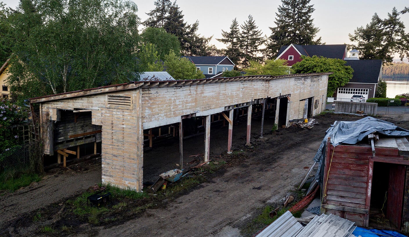 Abandoned building structure with wooden walls, partially raised on stilts, surrounded by trees and residential homes in the background.