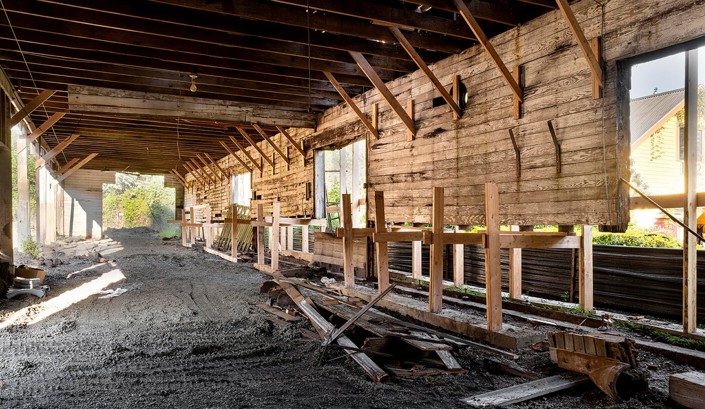 Interior of a construction site featuring exposed wooden beams, a dirt floor, and incomplete walls. Natural light filters through open windows.