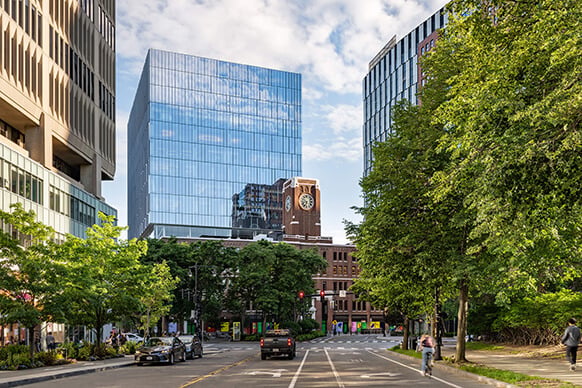 City street scene featuring glass office buildings and historic clock tower, with trees lining the road and vehicles and bicycles visible.