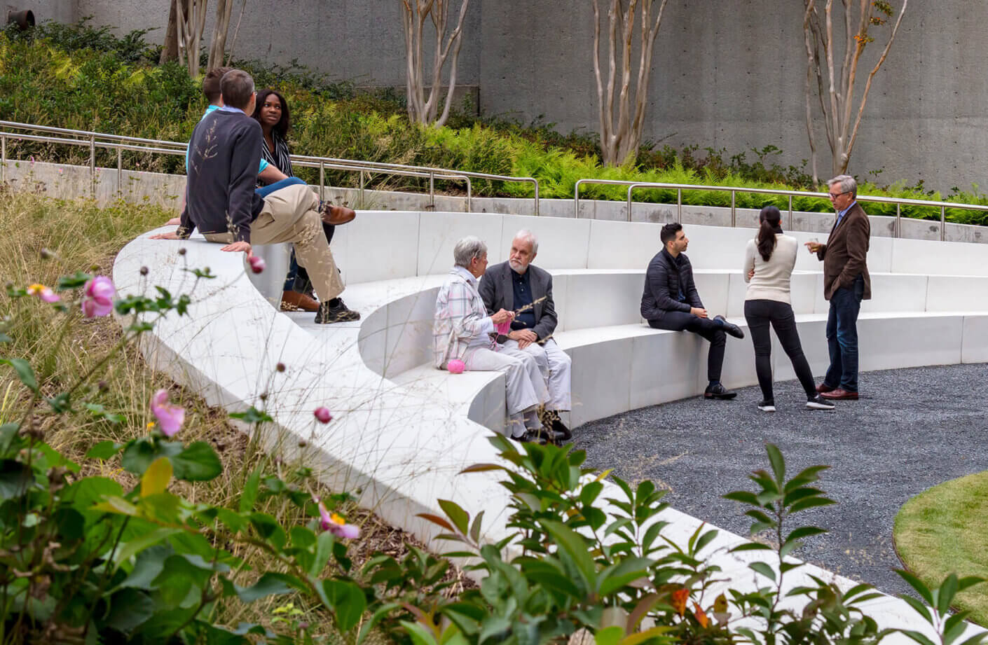 People sit and stand at the outer seating wall, with seasonal flowers in the foreground