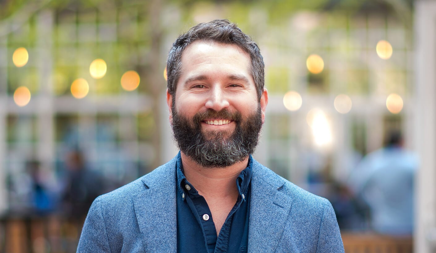 Headshot of Brad Rogers, wearing a blue jacket, outdoors with blurred lights behind.