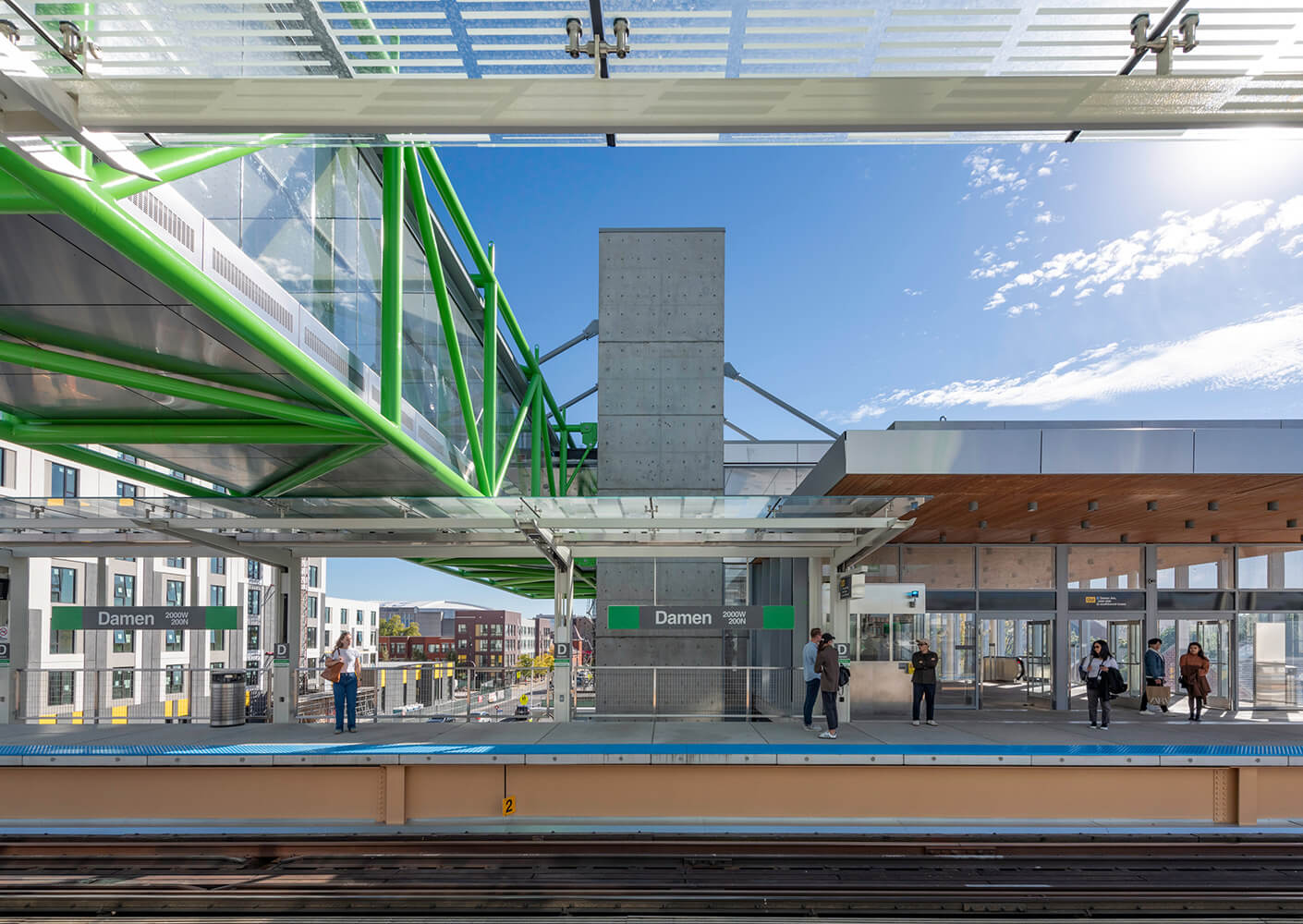 Station platform with green steel beams, and commuters on a sunny day.
