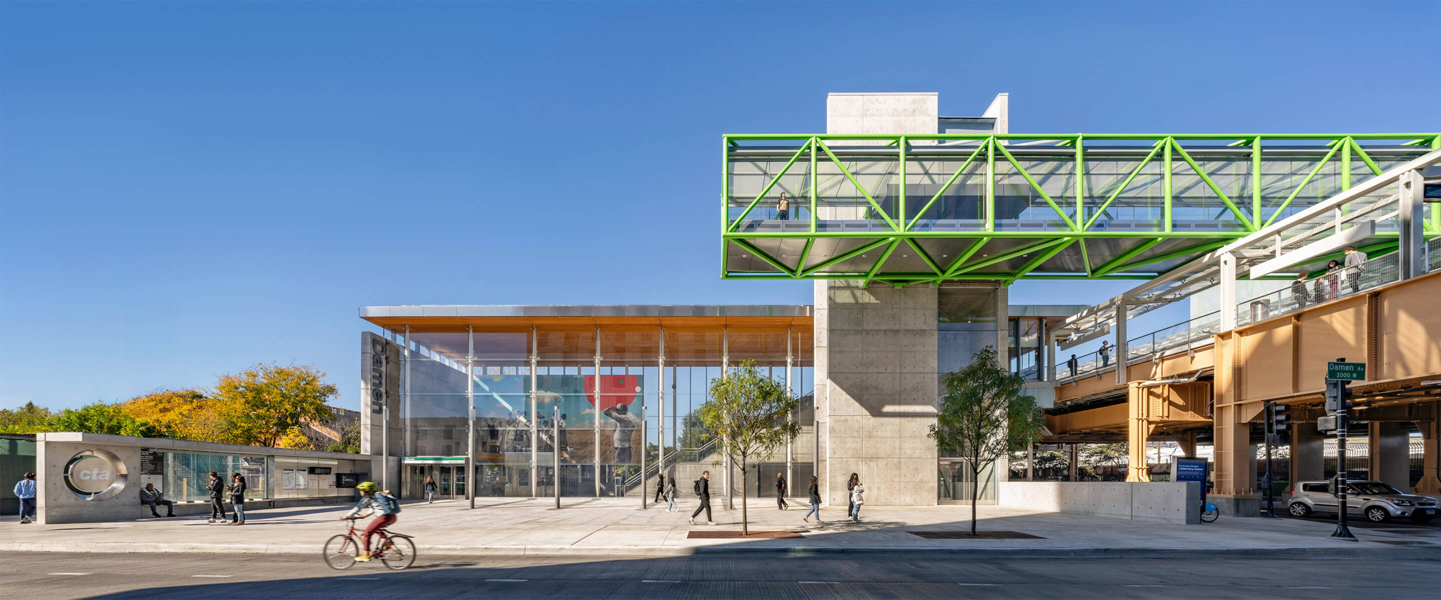 Modern train station with green skywalk, cyclists, and pedestrians under a clear blue sky.