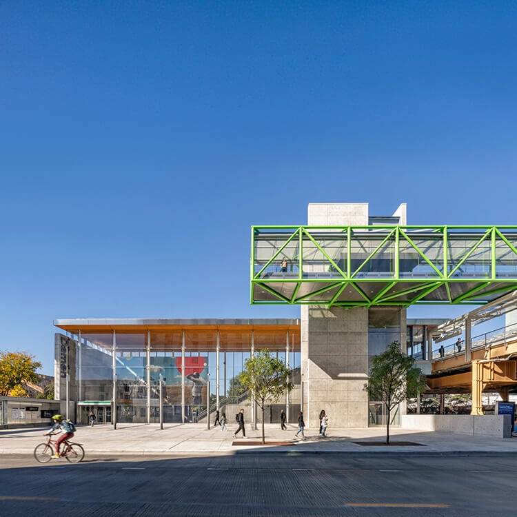 Modern train station with green skywalk, cyclists, and pedestrians under a clear blue sky.