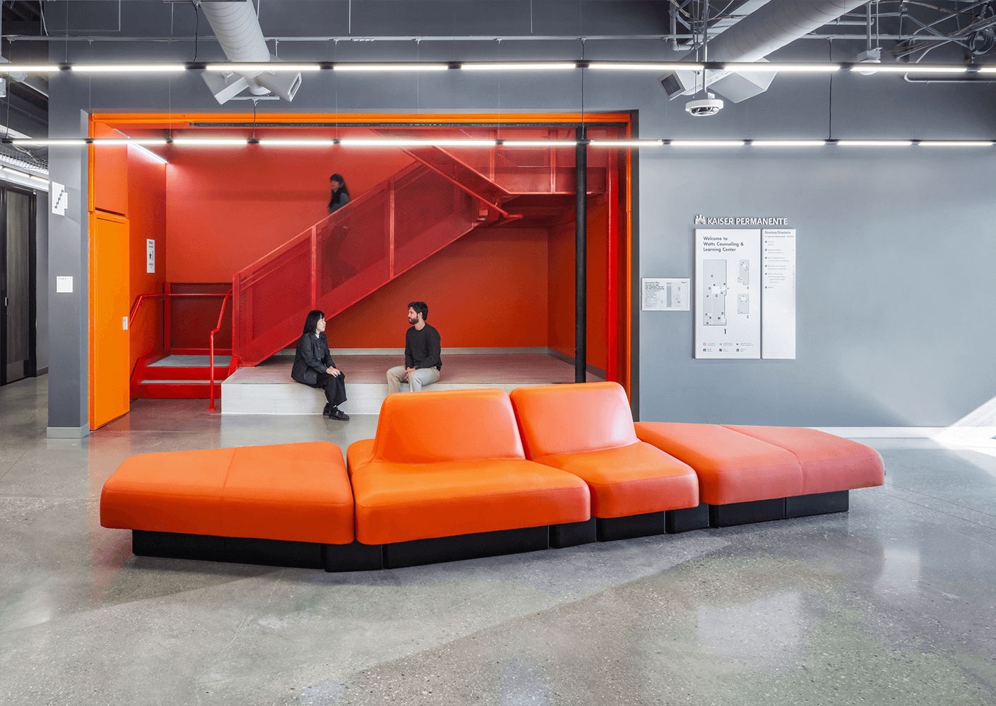 Modern office lobby with orange seating, red stairs, and people conversing and walking.