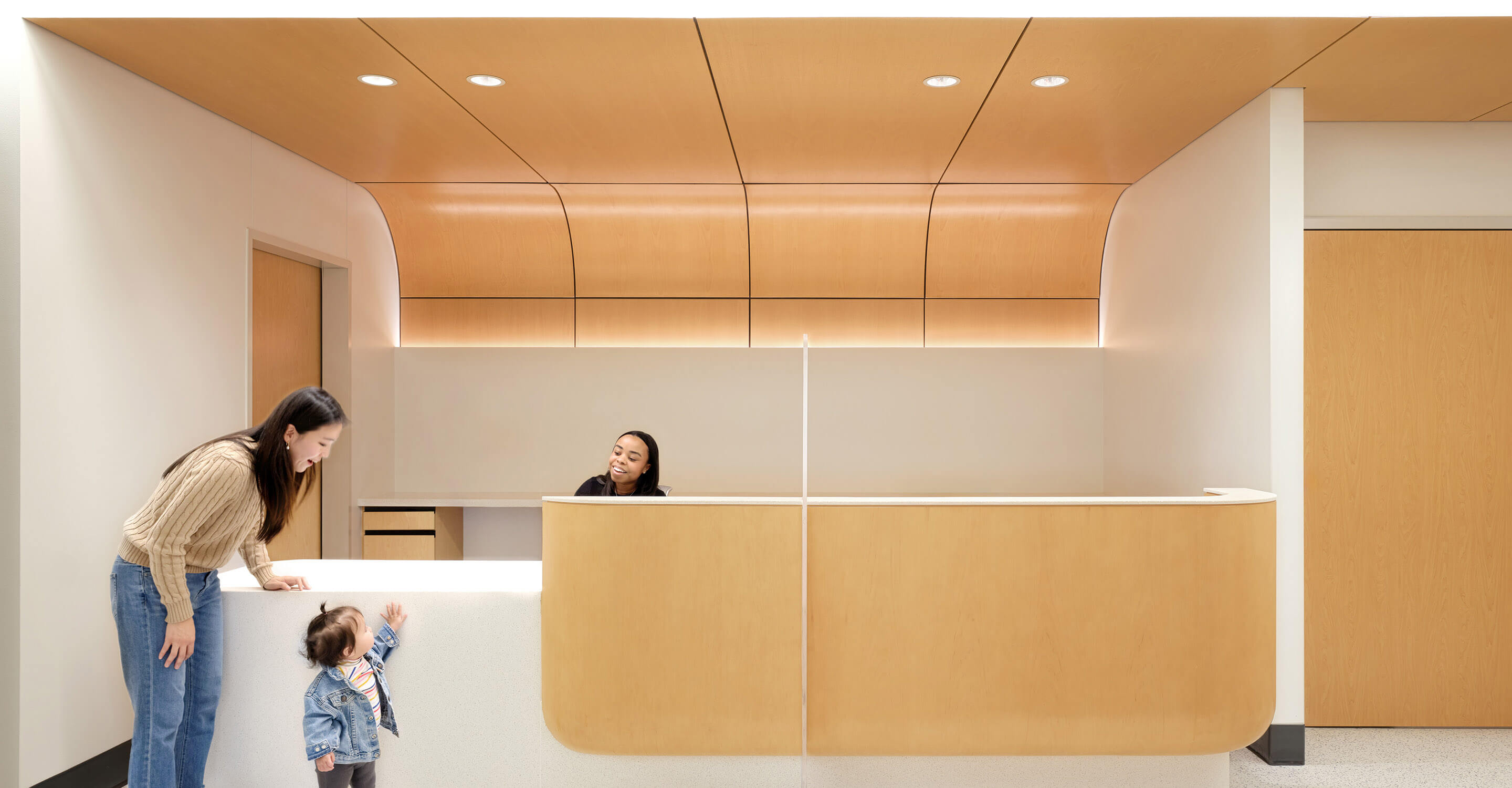Modern reception area with woman, child, and receptionist interacting, surrounded by warm wood panels