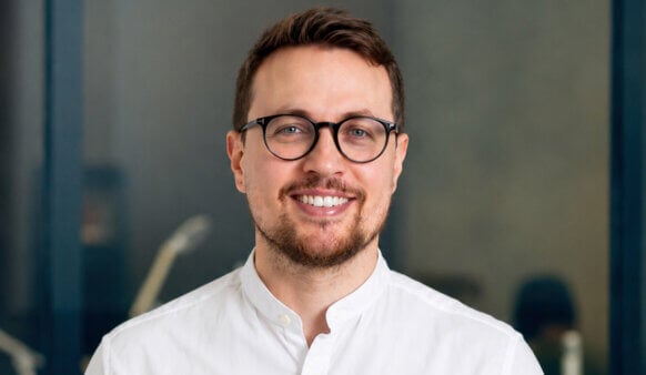Smiling man with glasses wearing a white shirt, posing in an office setting with a neutral background.