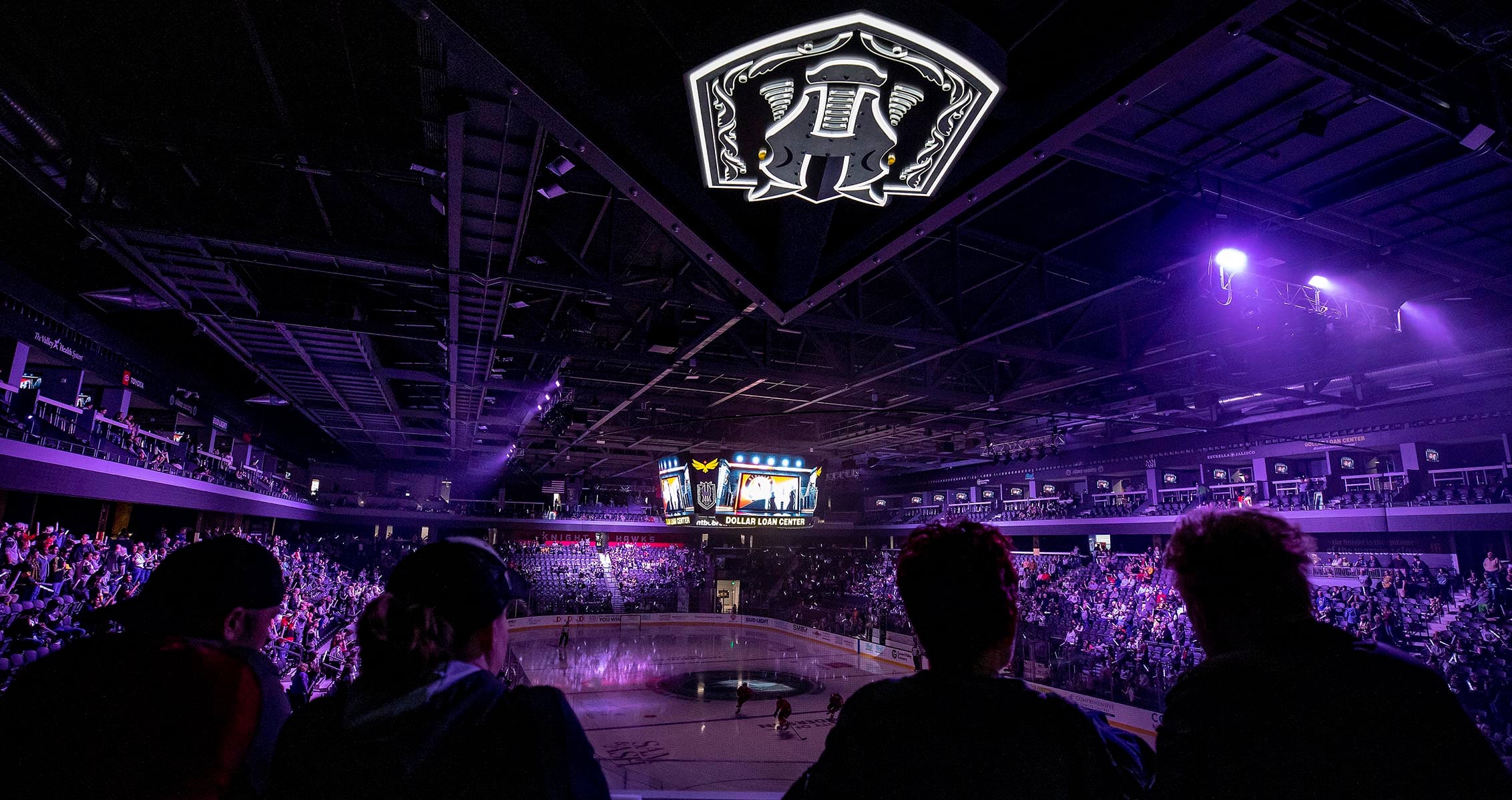Fans watch from the stands as a hockey game is about to begin at the arena, with purple lighting, a large center-hung scoreboard, and the team logo illuminated on the ceiling.