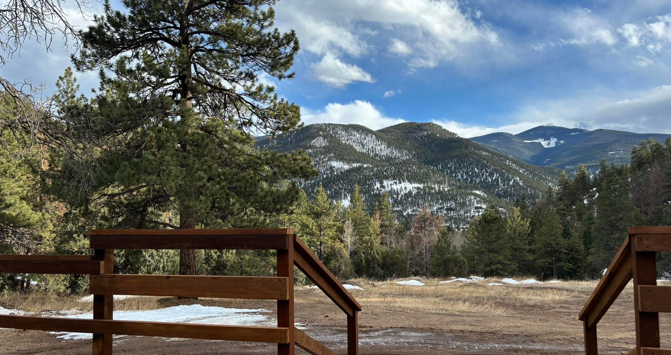View from a wooden deck looking out toward snow-dusted evergreen mountains under a partly cloudy blue sky, with tall pine trees in the foreground and patches of snow on the ground