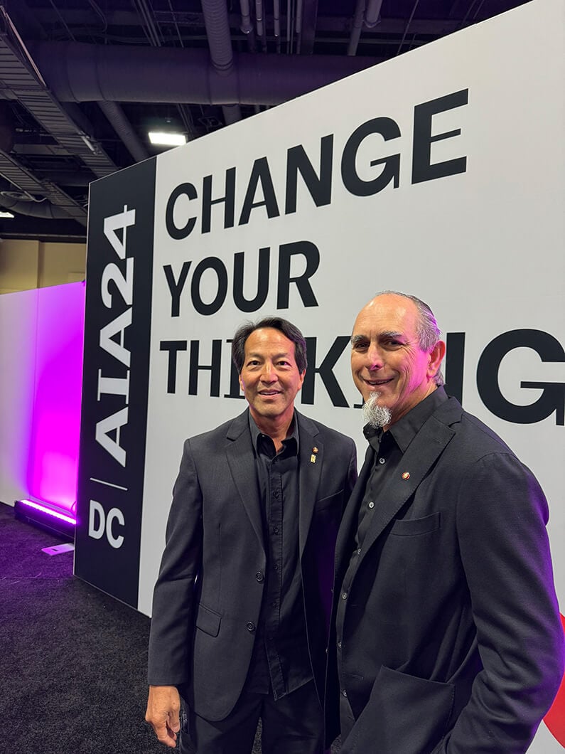 Two men in black suits at a conference with a CHANGE YOUR THINKING sign in the background.