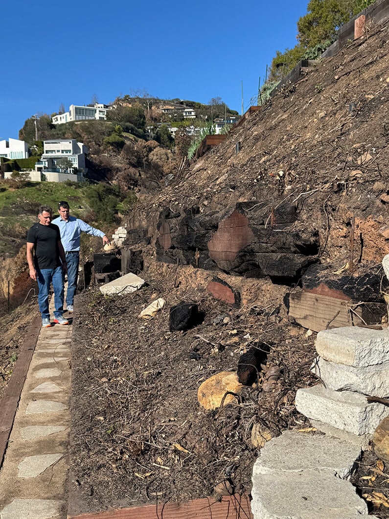 Two men examining a hillside with burnt vegetation, with houses visible in the background on a sunny day.