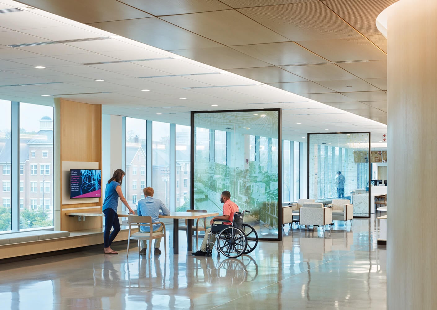 Modern hospital lounge with patients, wheelchair, and staff discussing care. Bright, spacious interior with large windows.