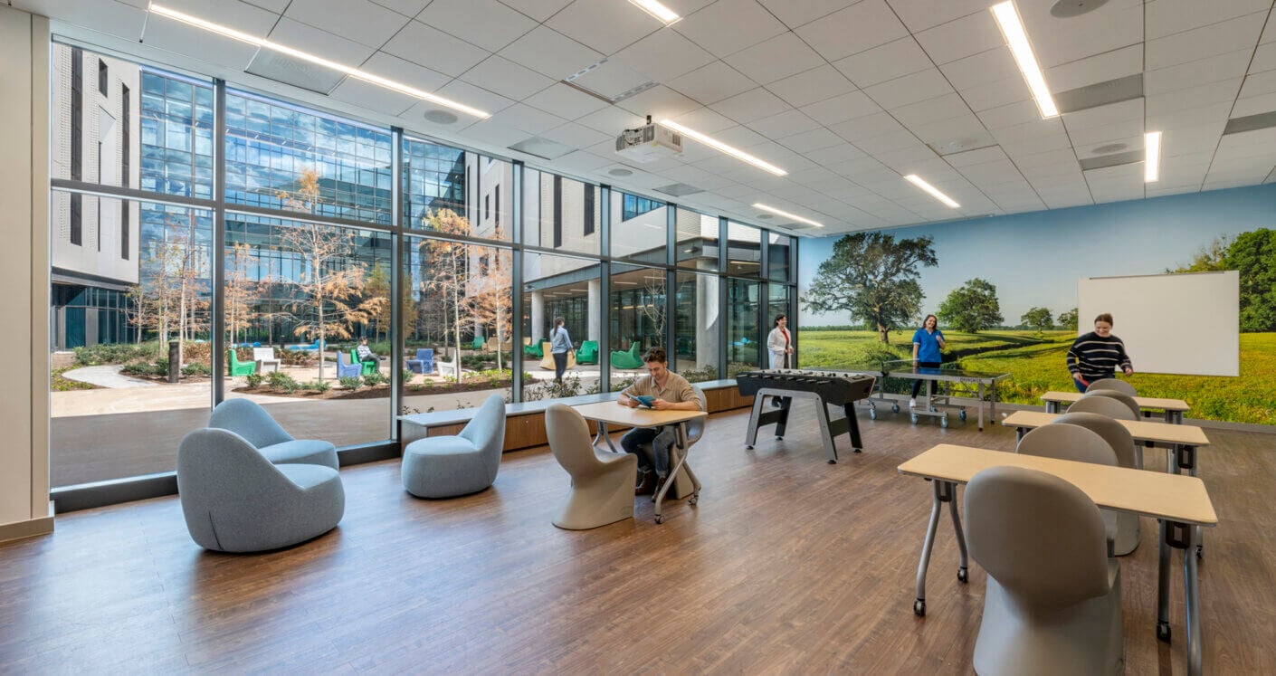 Modern recreational space with natural light, comfy seating, and wall mural, featuring people enjoying activities.