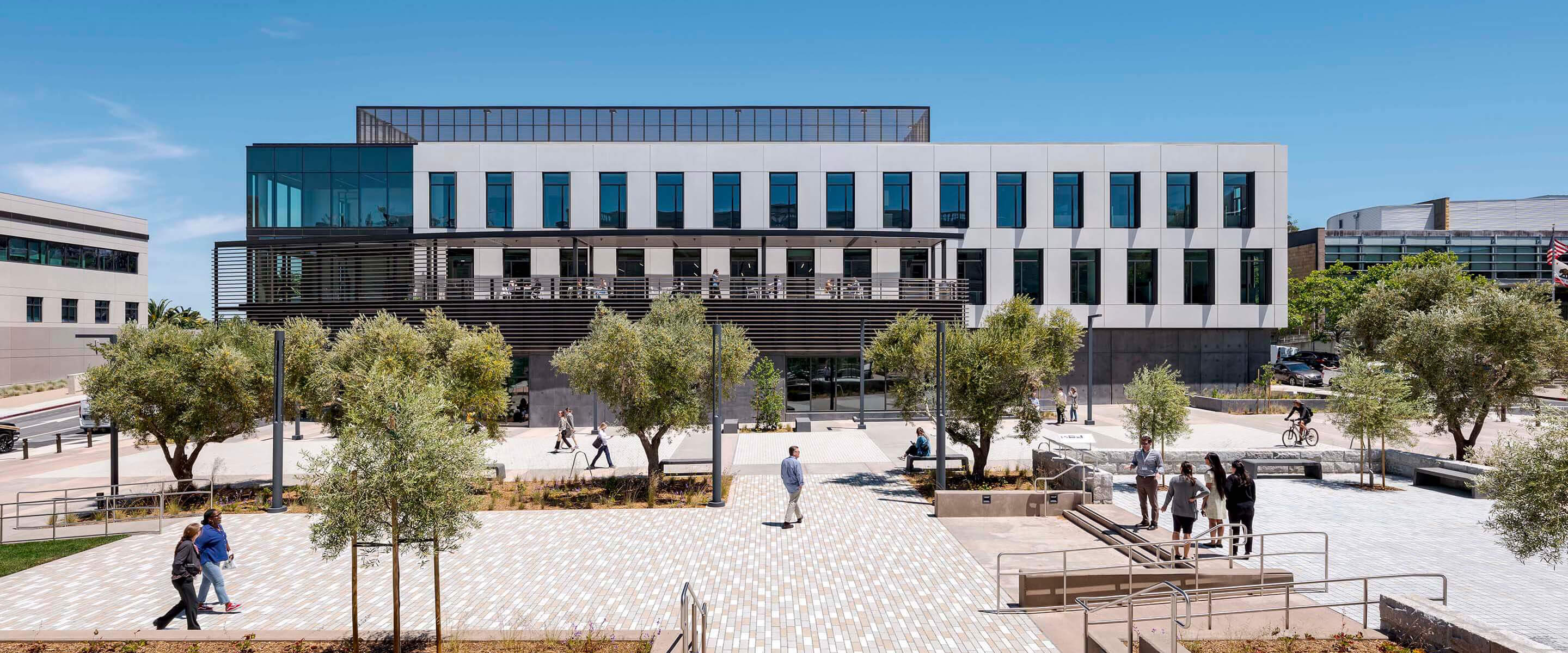 Modern office building with trees and people walking in a sunny plaza.
