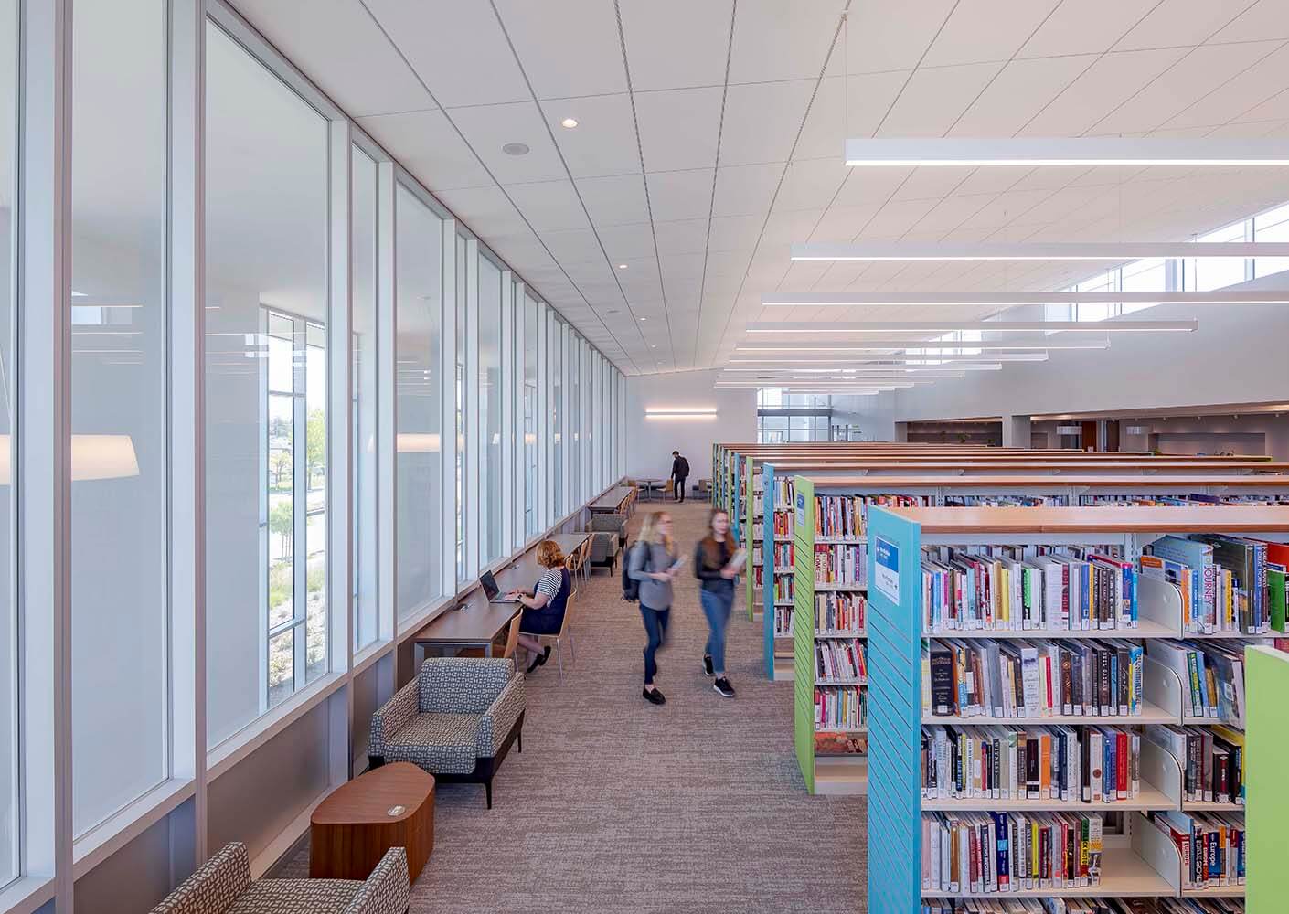 Modern library with bookshelves, large windows, and people studying.