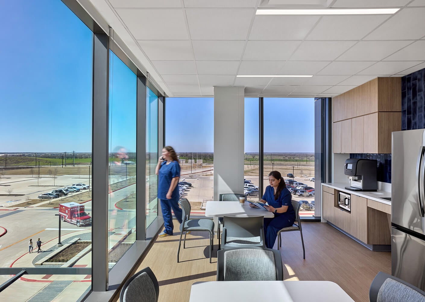 Hospital break room with two people in scrubs, large windows, and outdoor view.