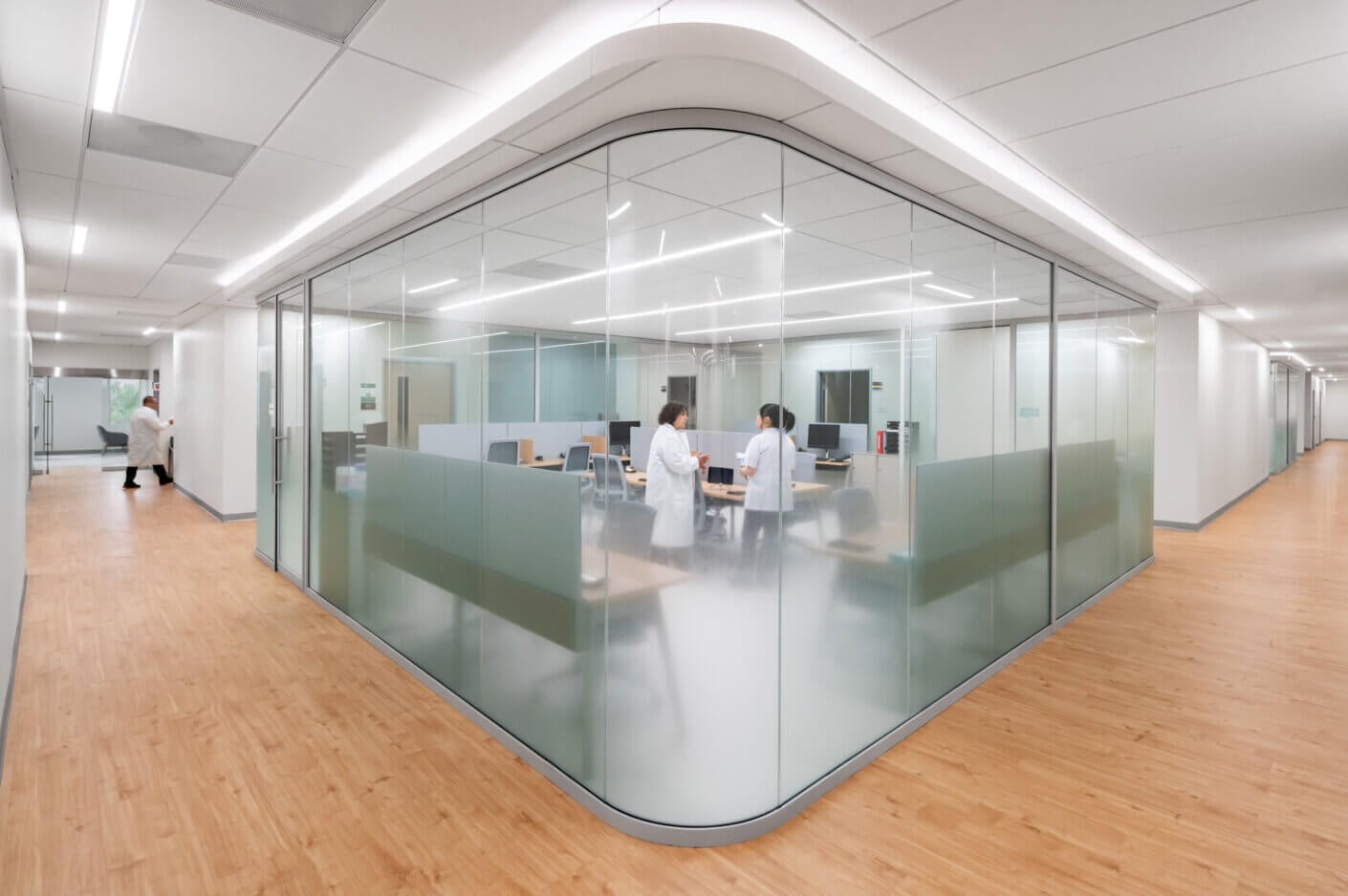 Modern office hallway with glass walls, two people in lab coats in a meeting.