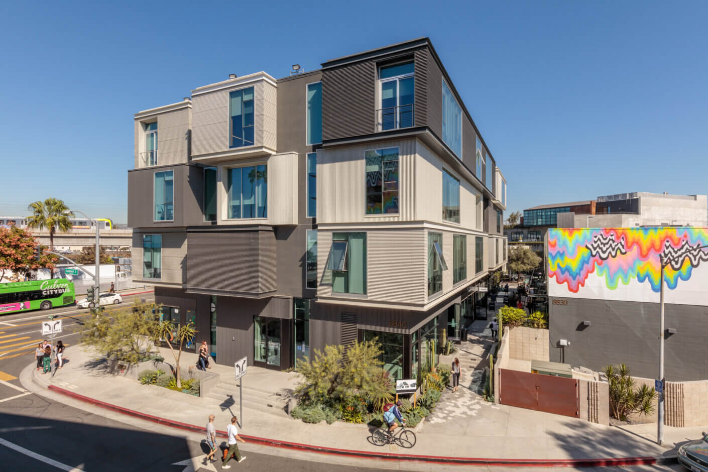 Modern urban building on sunny street with colorful mural and people walking.