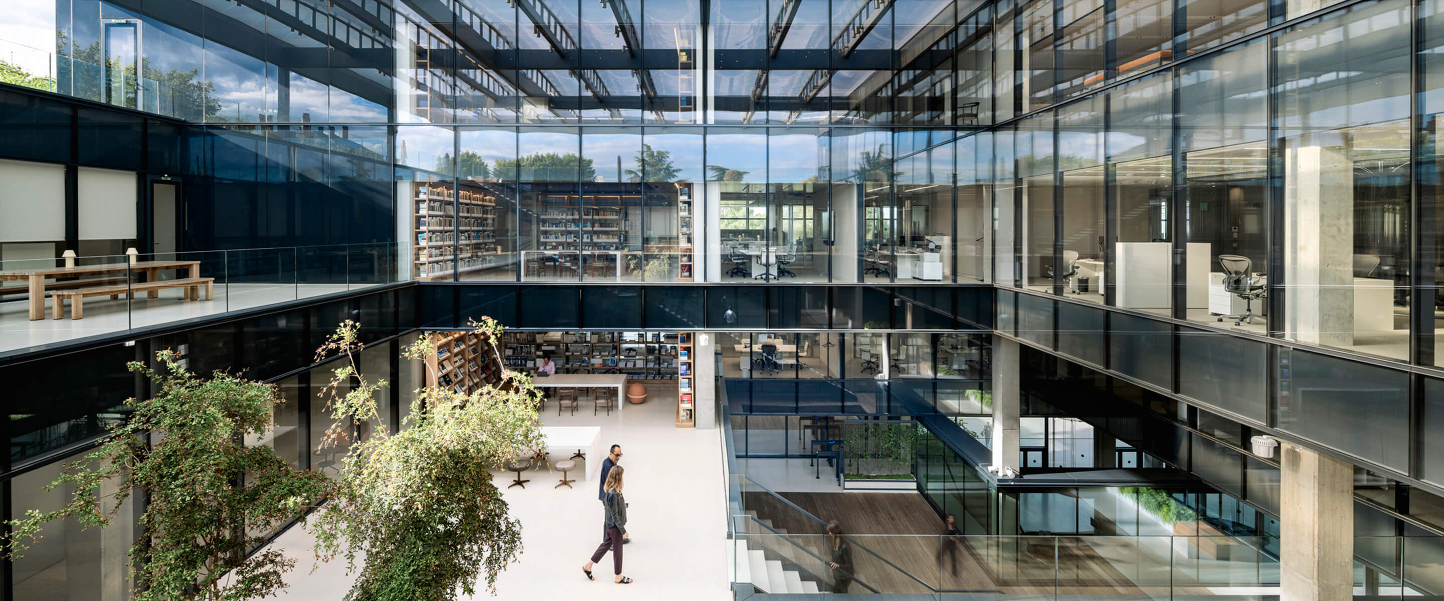 Modern glass office atrium with greenery and people walking.