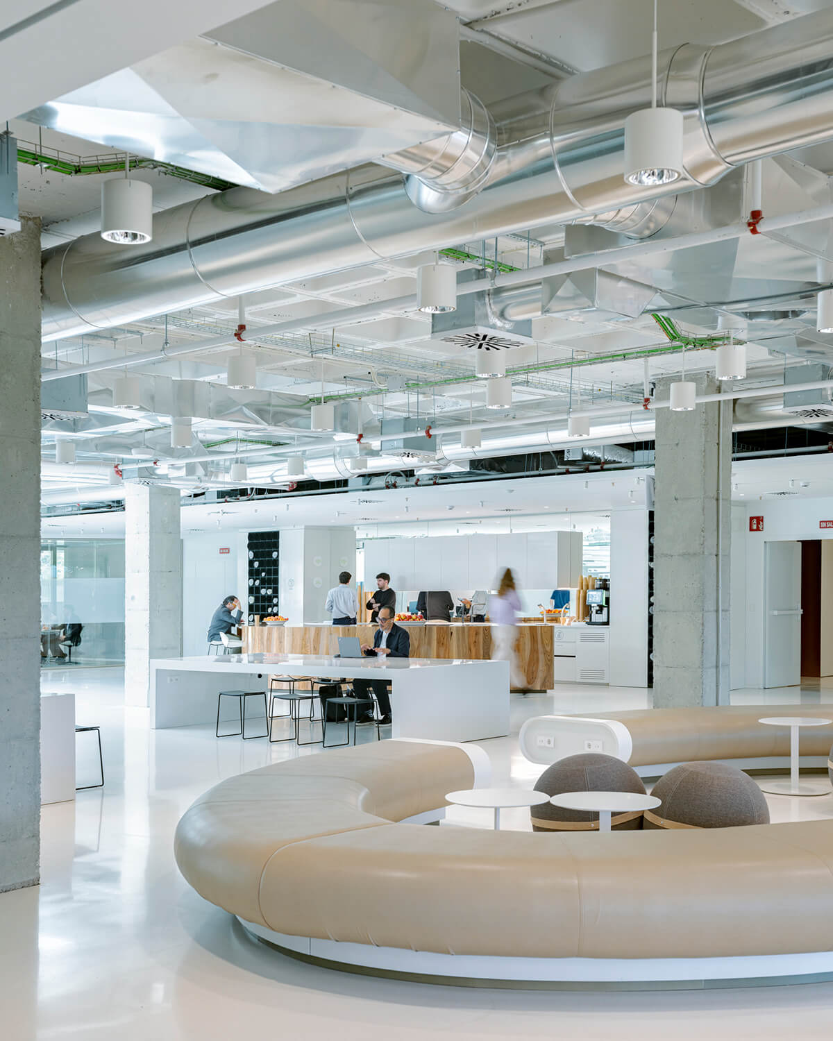 Modern office interior with industrial ceiling and collaborative workspace with curved seating and a dining area.