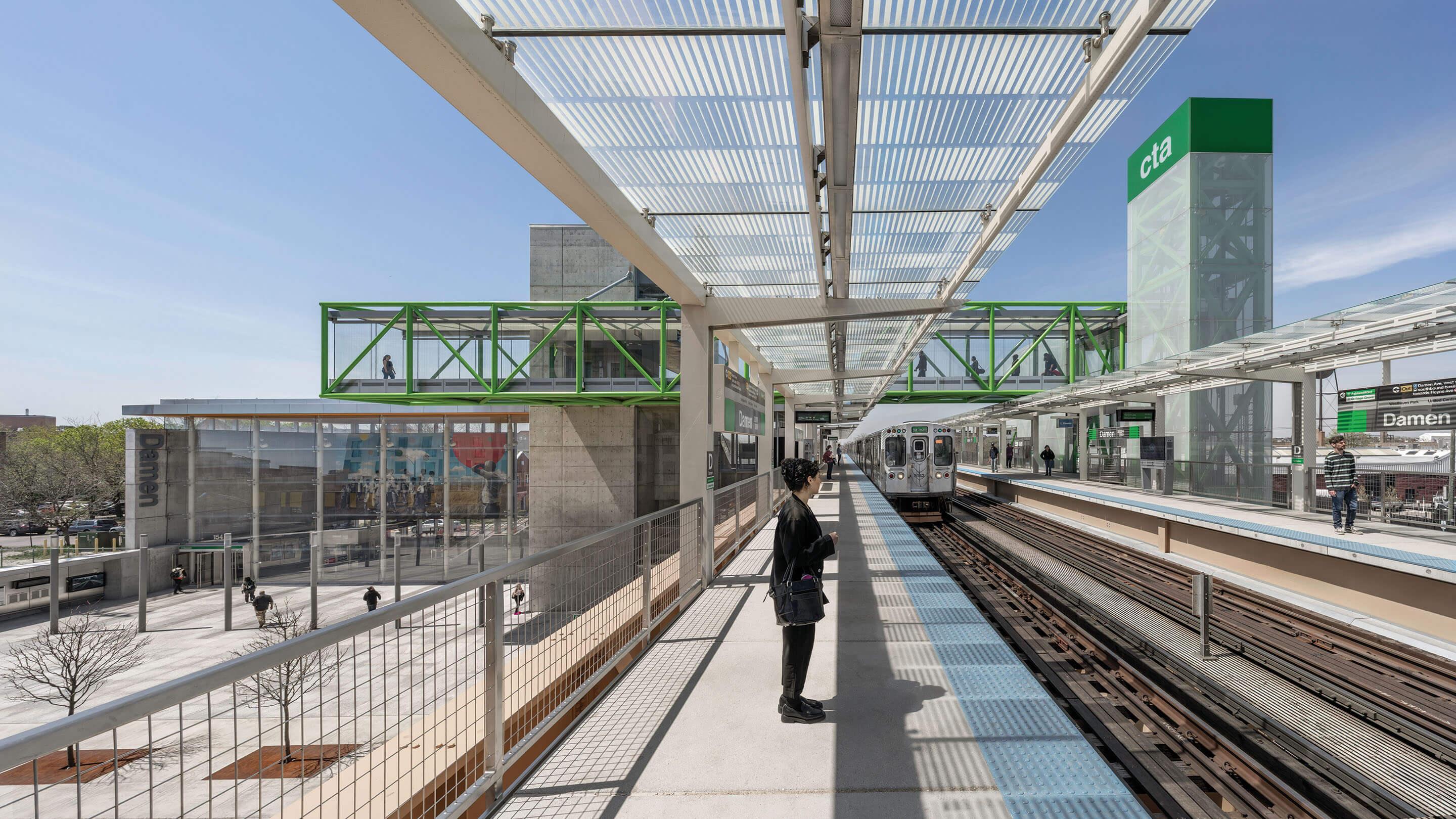 A person waits on the train platform as a train approaches with other commuters in the background