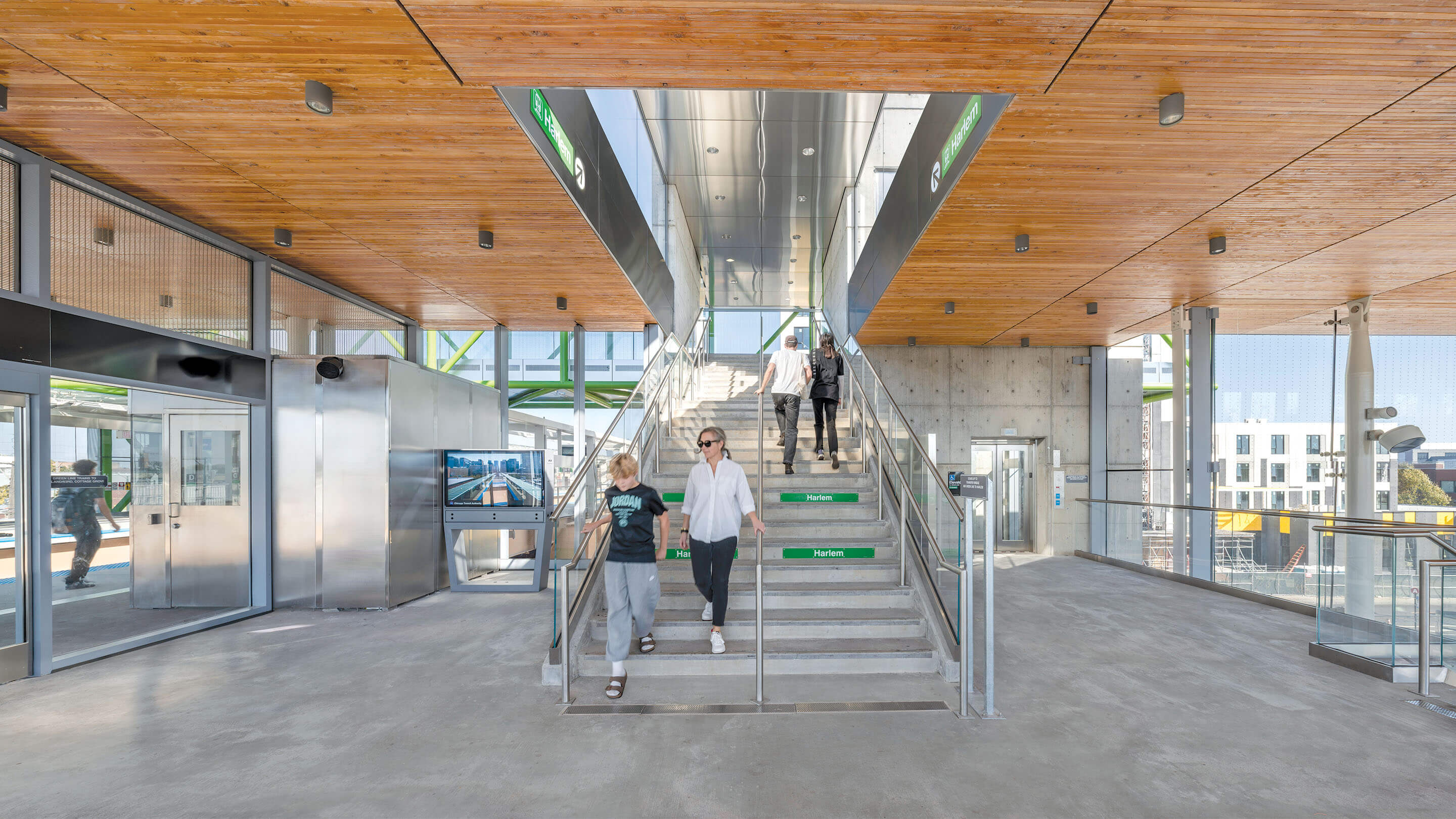 Passengers descend the stairs of a train station interior with a wood ceiling and green wayfinding signage.
