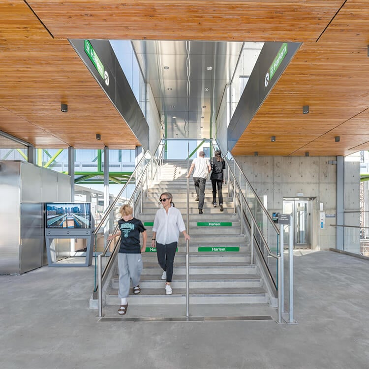 Passengers descend the stairs of a train station interior with a wood ceiling and green wayfinding signage.