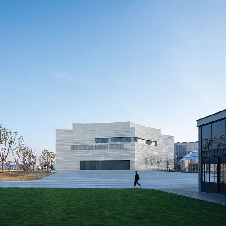 Modern white building with curves and angles, set behind a paved plaza and grassy area on a winter day.