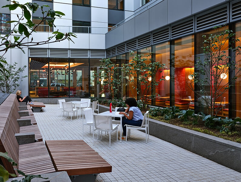 Outdoor terrace area with seating, plants, and people relaxing near a building.