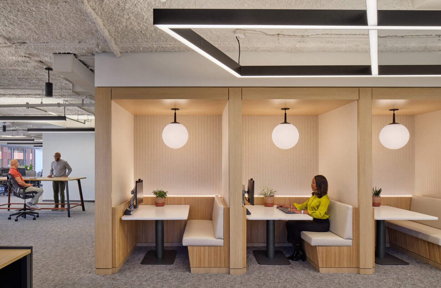 Office interior featuring collaborative workspaces with distinct seating areas and modern lighting, showcasing individuals engaged in work activities.