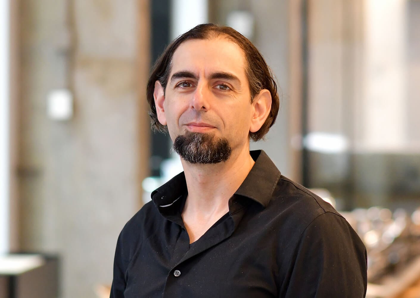 Portrait of a bearded man in a black shirt, smiling against a blurred background.