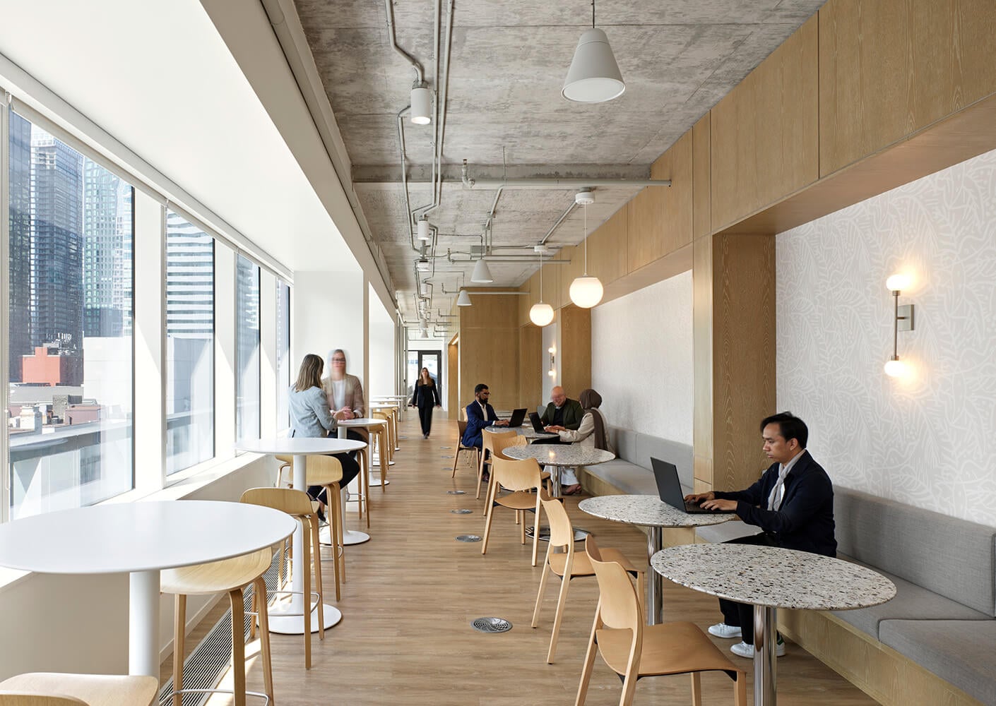 Bright workspace with large windows, wooden elements, and seating areas for collaboration and individual work. Professionals engaged in activities.