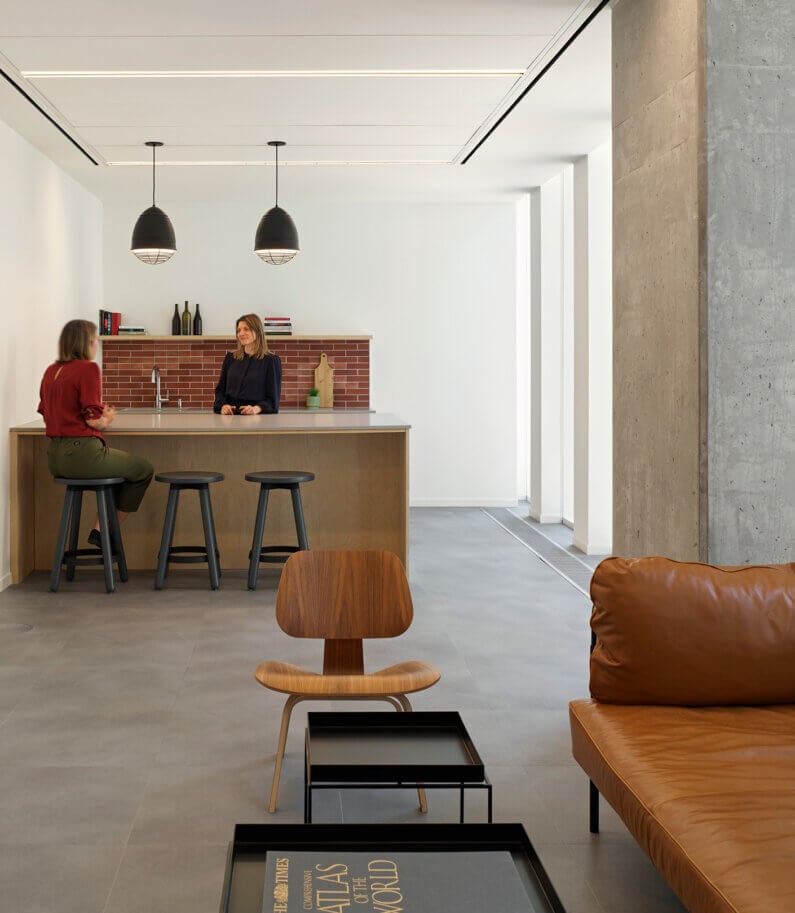 Two women converse at a kitchen island with wooden elements, black stools, and pendant lights. A brown leather sofa and wooden chair are visible in the foreground.